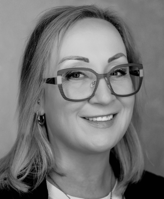 A person with glasses smiles warmly, looking towards the camera in a black and white portrait.