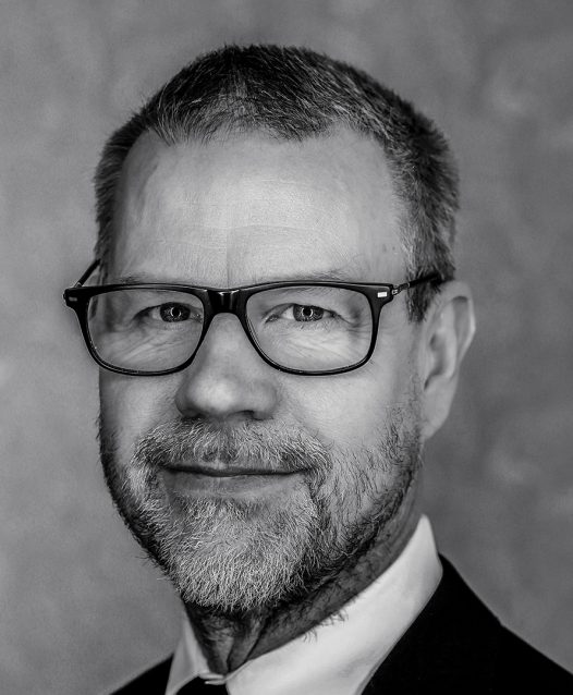 A man with glasses and a beard is wearing a suit and tie, looking directly into the camera with a neutral expression.