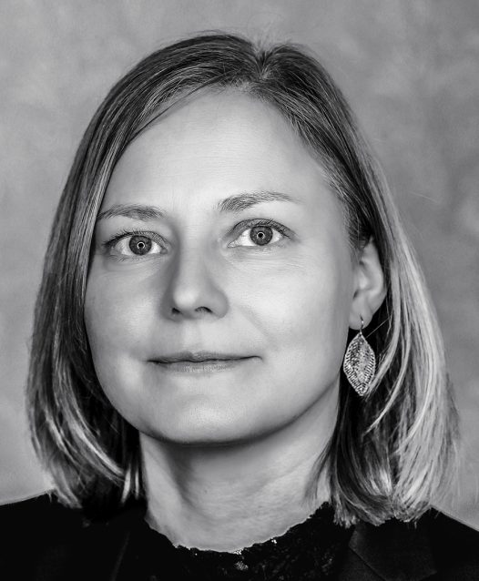 A grayscale portrait of a woman with shoulder-length hair, wearing a dark top and large, leaf-shaped earrings, looking directly at the camera.