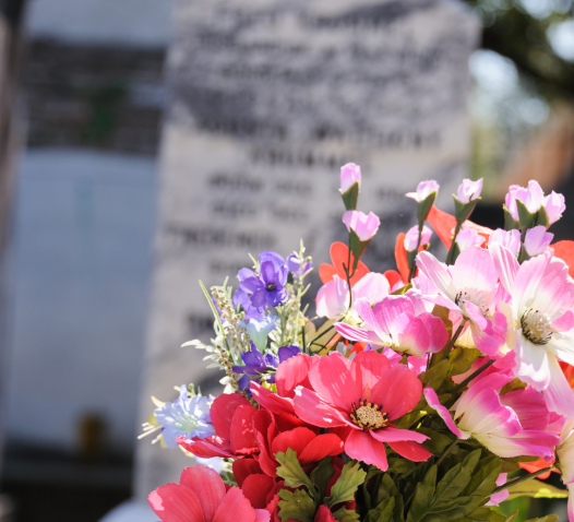 A bouquet of vibrant red, pink, and purple flowers is placed in front of a blurred gravestone in a cemetery.